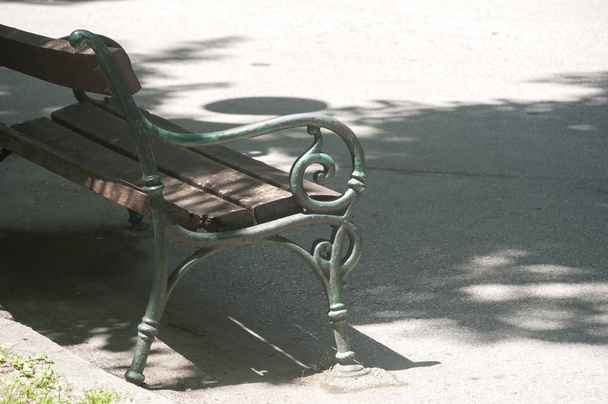 Banco de jardim vintage com construção de metal e assentos de placa de madeira no beco do parque de asfalto na sombra da árvore no dia ensolarado do verão
 - Foto, Imagem