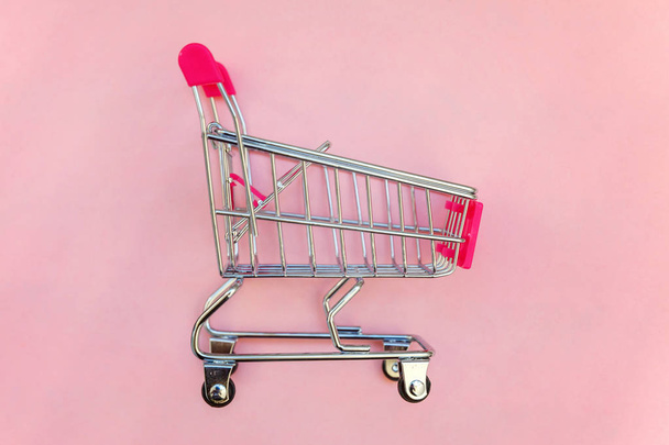 Small supermarket grocery push cart for shopping toy with wheels and pink plastic elements on pink pastel color paper flat lay background. Concept of shopping. Copy space for advertisement - Zdjęcie, obraz