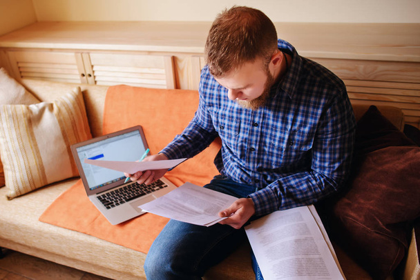 Nuori mies opiskelee kotoa istuu sohvalla tilalla asiakirjoja työskennellessään kannettavan tietokoneen
. - Valokuva, kuva