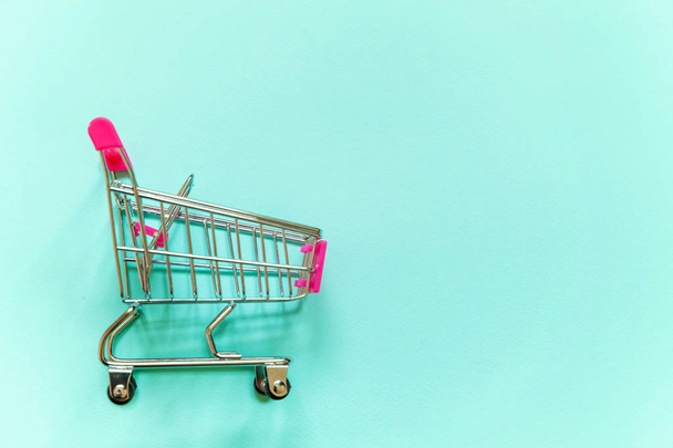 Small supermarket grocery push cart for shopping toy with wheels and pink plastic elements on blue pastel color paper geometric flat lay background. Concept of shopping. Copy space for advertisement - Fotoğraf, Görsel