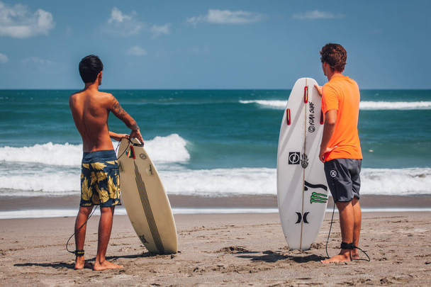 vista trasera de dos jóvenes de pie con tablas de surf en la playa de arena
 - Foto, imagen