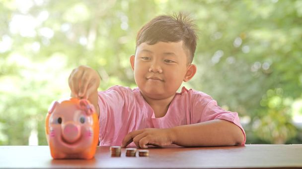 Ázsiai fiú, egy boldog mosollyal szórakoztató elhelyezés érméket a piggy bank, lassú mozgás. bokeh háttér. Pénzt takaríthatunk meg, és pénzt gyűjtött a jövő. - Fotó, kép