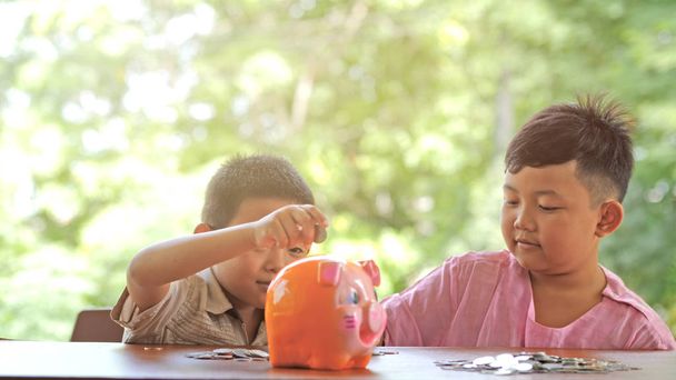 Menino asiático duas pessoas feliz sorriso divertido colocando moedas em um banco porquinho, Slow Motion. Bokeh background. Poupar dinheiro e recolher dinheiro para o futuro
. - Foto, Imagem