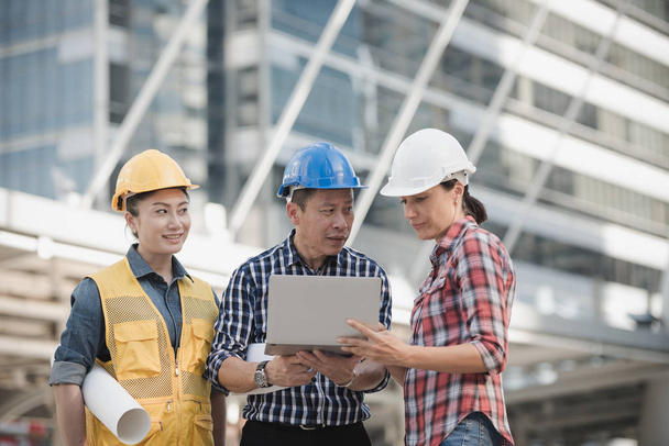 Die asiatische Ingenieurgruppe berät die Bauarbeiter vor Ort, während sie Laptop und Blaupause in der Hand hält. im Hintergrund der Stadt. Technik, Teamwork-Konzept - Foto, Bild