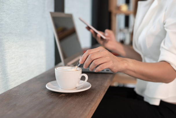 Молодая бизнесвумен держит ложку и помешивает горячий кофе на деревянном столе, а в кафе работает телефон с ноутбуком - Фото, изображение