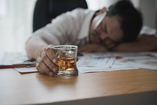 ウイスキーのグラスを持っているアジアのビジネスマンをストレス彼は眠っているとデータチャート、オフィスの机でビジネス文書。アルコール依存症-飲酒ビジネスマンの概念 - 写真・画像