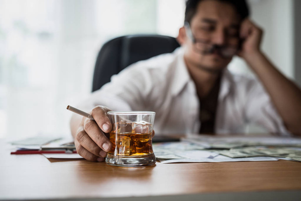 Gestresster asiatischer Geschäftsmann, der ein Glas Whiskey in der Hand hält und raucht. Er schläft auf dem Geld, Datentafeln, Geschäftsdokumenten am Schreibtisch. Alkoholsucht - Konzept betrunkener Geschäftsleute - Foto, Bild