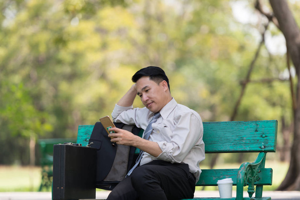 仕事の後、公園でリラックスしたアジア人ビジネスマン。彼は晴れた日には木の影を楽しむ電話を探している。起業家の救済や休憩時間の概念 - 写真・画像
