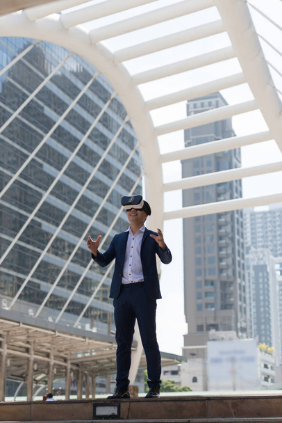 Jeune homme d'affaires asiatique en costume et lunettes de réalité virtuelle qui essaie de toucher quelque chose de sa main avec l'arrière-plan de la ville. Technologie conceptuelle et activités de simulation - Photo, image