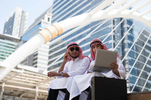 Dva mladí arabští podnikatelé sedí mluvit při práci s notebookem pozadí bangkok města, obchodní čtvrti s vysokou budovou oslnil - Fotografie, Obrázek