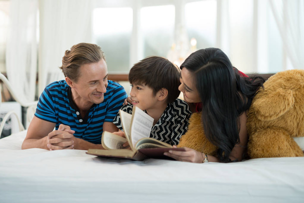 Ο μπαμπάς και η μαμά κοιτάζοντας το γιο μου διαβάζοντας ένα βιβλίο είναι στο κρεβάτι στις ιδέες της οικογενειακής εκπαίδευσης. - Φωτογραφία, εικόνα
