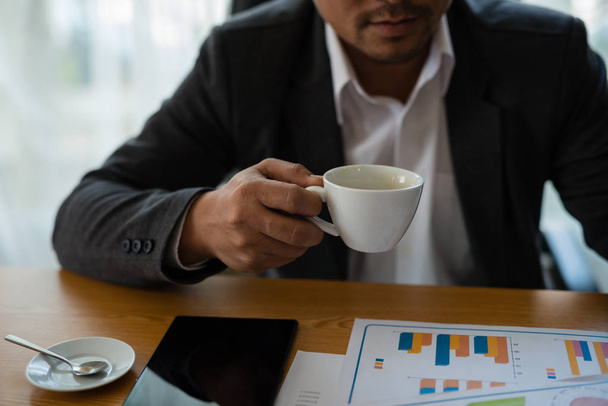 Young businessman holding a coffee cup in hand, looking at the financial statements of the Company in the document shown as a graph. Concept Marketing Investment - Photo, Image
