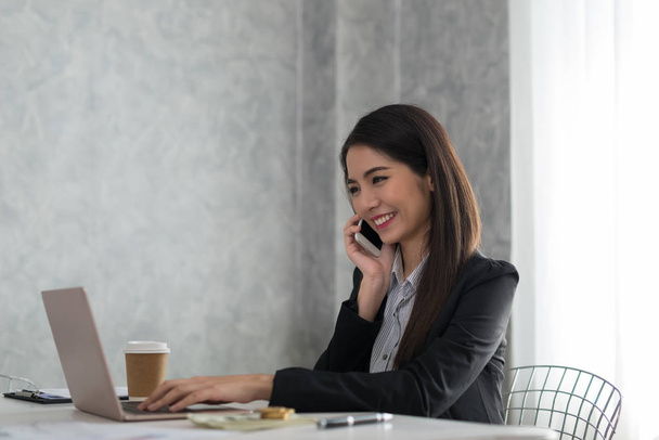 Красива азіатська молода бізнес-леді працює на ноутбуці, поки посміхається телефон у своїй робочій станції за столом з яскравим вікном у фоновому режимі
 - Фото, зображення