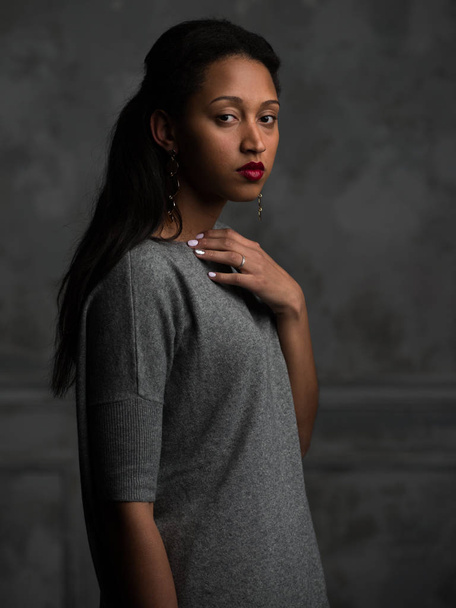 Side view of attractive young African American female in beautiful grey dress touching neck and looking at camera wistfully on grey background - Zdjęcie, obraz