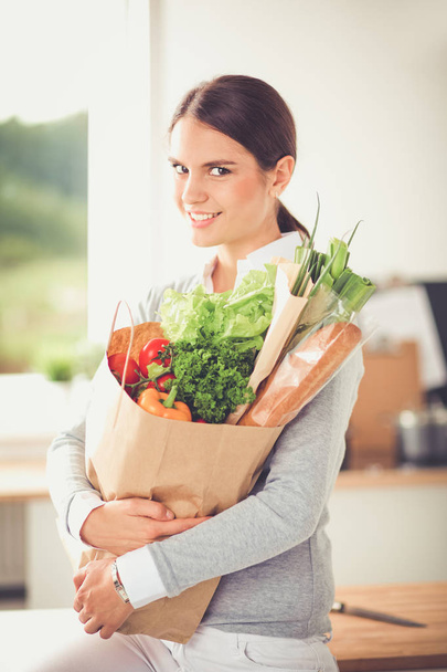 野菜の入った食料品の買い物袋を持っている若い女性。台所に立つ - 写真・画像