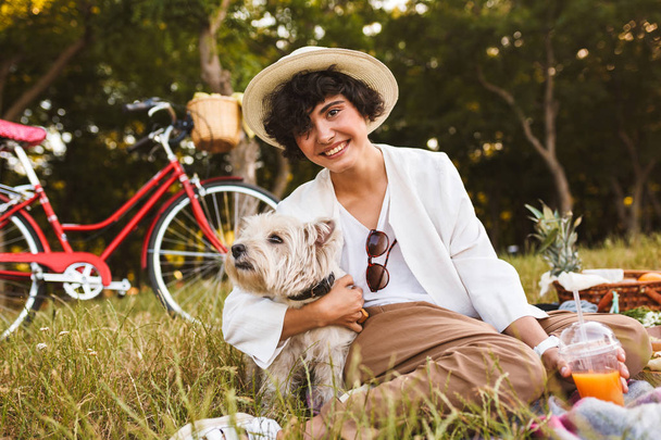 Όμορφο κορίτσι με το καπέλο που αγκαλιάζει μικρό χαριτωμένο σκυλί joyfullylooking στην κάμερα να ξοδέψετε χρόνο για πικνίκ στο πάρκο με κόκκινο ποδήλατο σε φόντο - Φωτογραφία, εικόνα