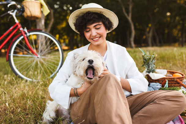 Красива дівчина в капелюсі тримає милий собаку радісно проводить час на пікніку в парку з велосипедом на фоні
 - Фото, зображення