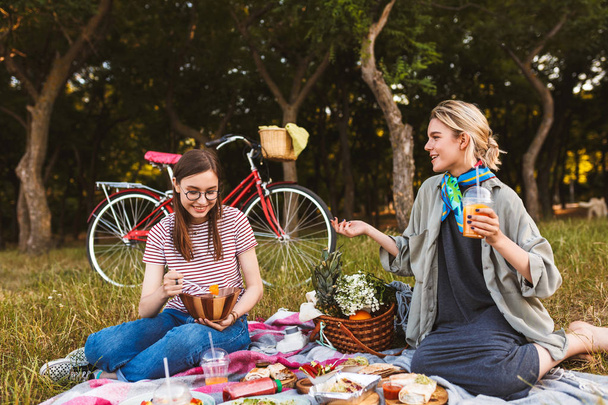 Hermosa chica sentada en una manta de picnic sosteniendo el jugo de naranja en la mano, mientras que la chica en camiseta rayada comiendo ensalada cerca de picnin en el parque
 - Foto, imagen