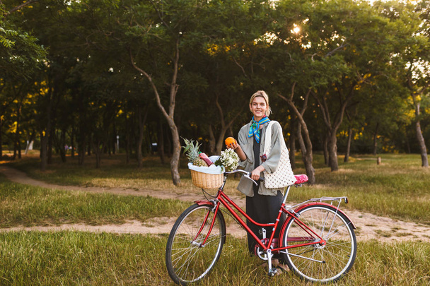 Hymyilevä tyttö punaisella polkupyörällä ja korilla täynnä villikukkia ja hedelmiä onnellisesti katsoen kameraan pitäen oranssia kädessä puistossa.
 - Valokuva, kuva