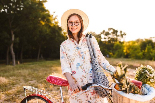 Cute smiling girl in eyeglasses on bicycle with basket full of fruits and wildflowers happily looking in camera in park - Photo, image