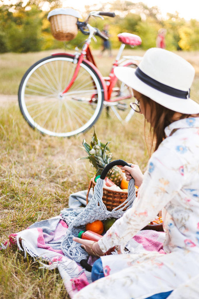 Panienka w biały kapelusz siedzi na piknik koc gospodarstwa butelka soku i torba z owocami, spędzać czas w parku - Zdjęcie, obraz