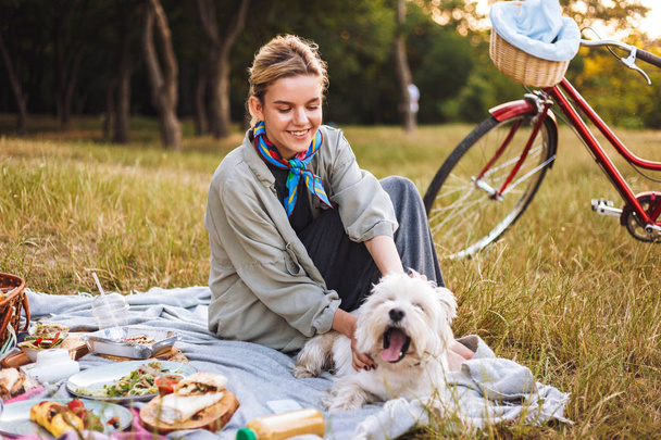 Nice smiling girl with cute dog happily spending time on beautiful picnic in park with red bicycle on background - Fotoğraf, Görsel