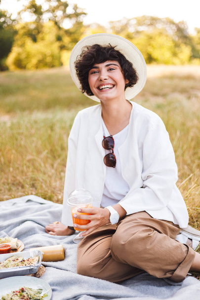 Beautiful smiling girl sitting in hat and white shirt holding cup with juice to go in hand happily looking in camera on picnic in park - Photo, Image