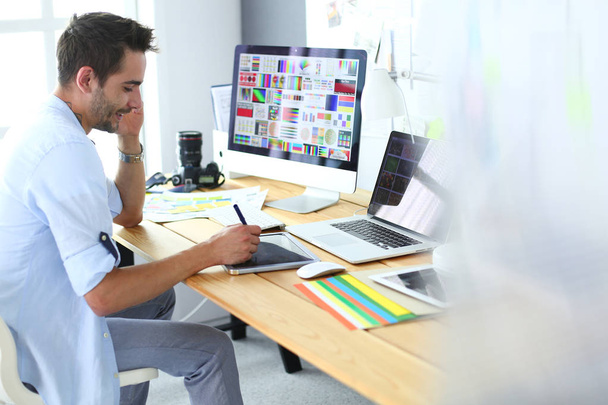 Retrato del joven diseñador sentado en el estudio gráfico frente a la computadora portátil y el ordenador mientras trabaja en línea. - Foto, imagen