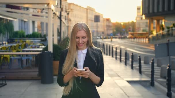 Formal business woman walking on street. Elegant blond woman in suit and walking on street and browsing smartphone with smile against urban background. - Footage, Video