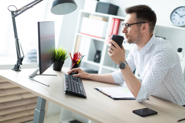 Un joven con gafas se para cerca de una mesa en la oficina, tiene un vaso de café en la mano y trabaja con una computadora. - Foto, Imagen