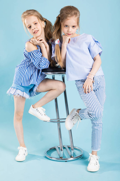 Mädchen-Zwillinge in hellblauer Kleidung posieren in der Nähe eines Barhockers auf blauem Hintergrund. - Foto, Bild
