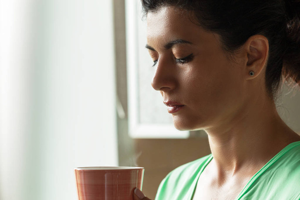A calm morning - a middle-aged woman drinks fresh hot coffee in a mug on the window background. - Zdjęcie, obraz