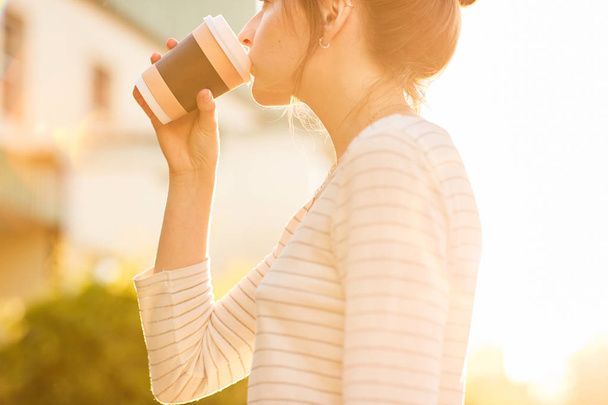 young woman drinking coffee from paper cup at sunset - Фото, изображение