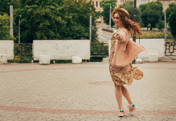Smiling happy girl in dress and hat on a city street and looking at camera. in a hat, a dress in a flower and a jacket of warm colors - Zdjęcie, obraz