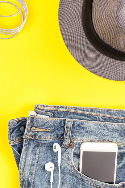 Flat lay of blue jeans, grey hat and smartphone headphones on bold yellow paper background with copy space. Overhead view of woman casual outfit. Trendy hipster look top view - Φωτογραφία, εικόνα