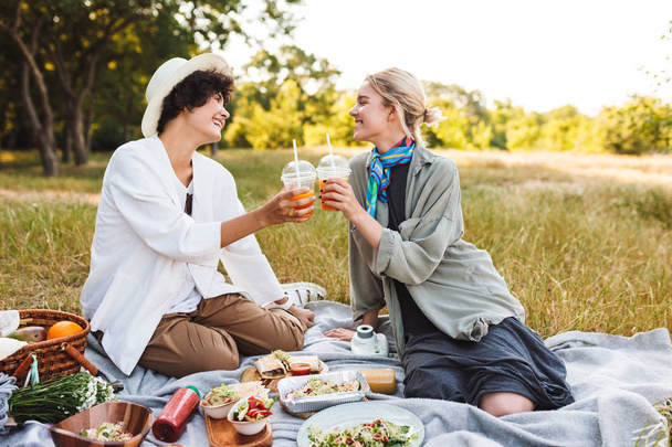 Две красивые девушки сидят на одеяле для пикника, держа в руках апельсиновый сок, и с удовольствием проводят время на пикнике в парке.
 - Фото, изображение
