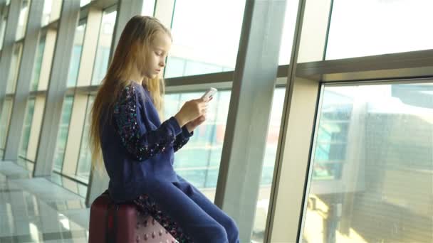 Adorable niña en el aeropuerto cerca de una gran ventana jugando con su teléfono
 - Imágenes, Vídeo