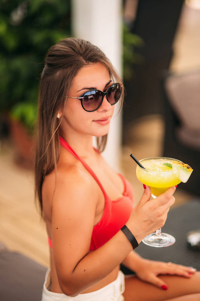 Mulher bonita vestindo maiô bebendo um coquetel colorido sentado em uma cabine do bar do clube de praia. Menina impressionante desfrutando de sua bebida na hora de verão
 - Foto, Imagem