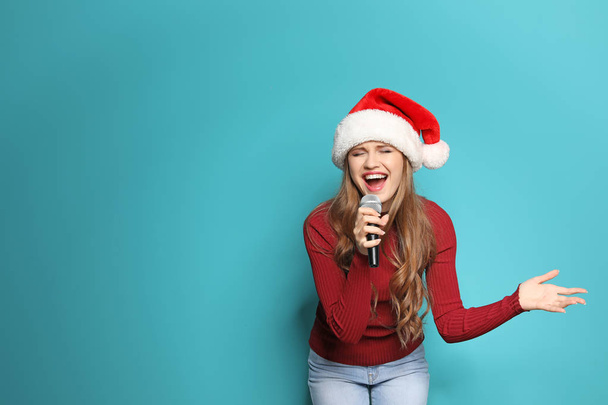 Young woman in Santa hat singing into microphone on color background. Christmas music - Fotografie, Obrázek