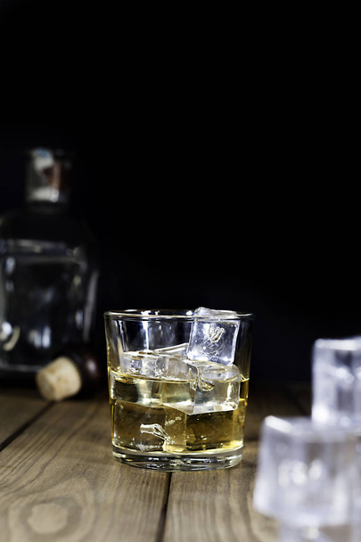 whisky de cristal con hielo, cubitos de hielo y botella sobre mesa de madera y fondo negro
 - Foto, imagen