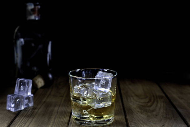 скляне віскі з льодом, кубиками льоду та пляшкою на дерев'яному столі та чорному фоні
 - Фото, зображення
