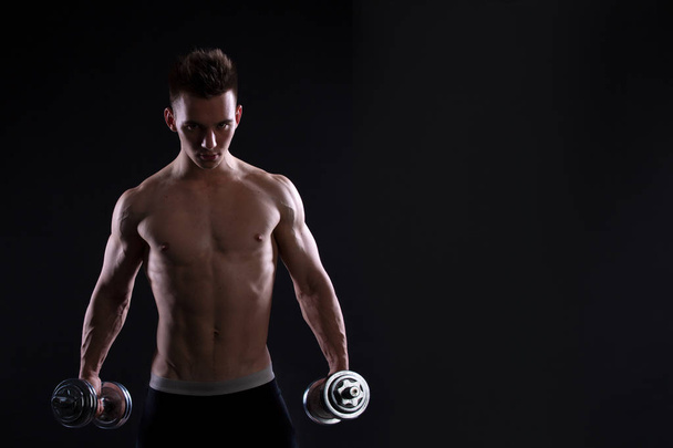 Αθλητική νεαρό άνδρα με έναν αλτήρα σε μαύρο φόντο. Γυμνό κορμό, μυώδες σώμα. Ισχυρή μύες στο στήθος και τους ώμους. Στούντιο shot, χαμηλών τόνων. Bodybuilding έννοια, αντίγραφο χώρου - Φωτογραφία, εικόνα