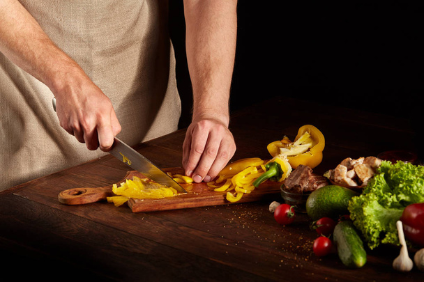 Chefs hands cutting yellow bell pepper for colorful barbecue composition on wooden table, close-up, selective focus. - Foto, imagen