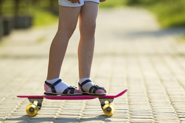 Girl slim legs in white socks and black sandals standing on pavement on plastic pink skateboard on bright sunny summer blurred background. Outdoors activities and healthy lifestyle concept. - Foto, afbeelding