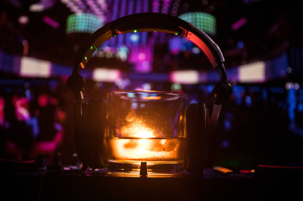 Glas met whisky met ice cube binnen op dj-controller bij nightclub. DJ koptelefoon op een glas van whilsky op muziek feest in nachtclub met disco lichten. Selectieve aandacht - Foto, afbeelding