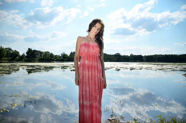 Young female model in outdoor photo session in Poland. Girl with long red dress stands near lake. - Photo, Image