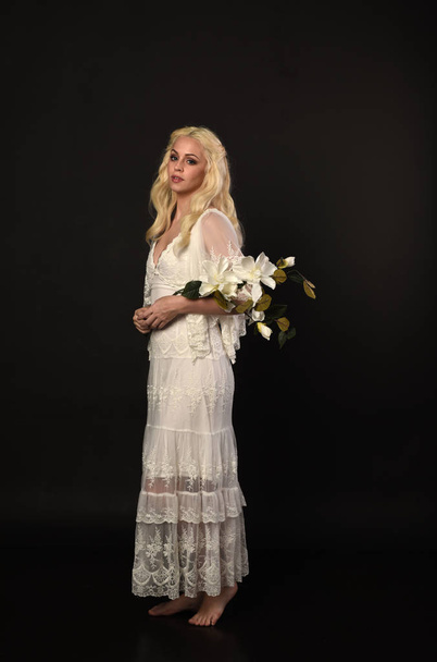 Pełna długość Portret Blondynka dziewczyna ubrana w białej sukni koronki. Stojący poza, gospodarstwa kwiaty. czarny studio tło. - Zdjęcie, obraz