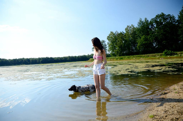 Jovem modelo feminino em sessão de fotos ao ar livre na Polônia. Menina de calças curtas e top de biquíni caminha no lago com seu cachorro
. - Foto, Imagem