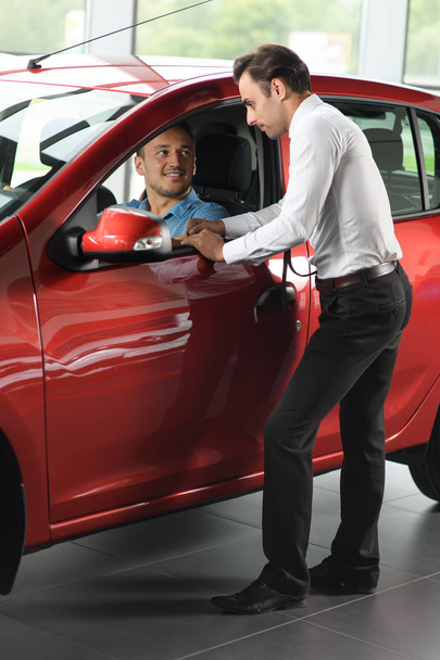 Πωλητής δείχνει επιλογές αυτοκινήτων για υπολογιστή-πελάτη. Πελάτης που κάθεται μέσα στο αυτοκίνητο. Το αυτοκίνητο είναι κόκκινο. Βρίσκονται σε αντιπροσωπεία αυτοκινήτων. - Φωτογραφία, εικόνα