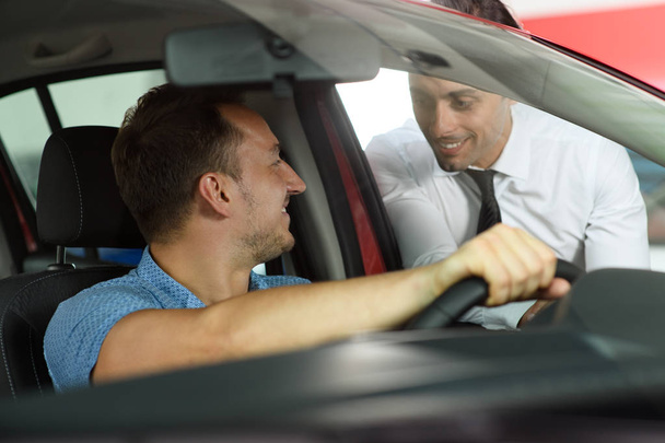 Salesman shows options for client. Client sitting in the car. Car is red. They are in car dealership. - Photo, Image